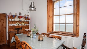 Une salle à manger avec une table en bois, des chaises, une fenêtre donnant sur la mer et une étagère où sont disposés de la vaisselle et des objets de décoration.
