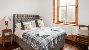 Une chambre à coucher avec une tête de lit capitonnée, une couvre-lit à motifs, une fenêtre avec vue, une table de chevet avec une lampe et un fauteuil.