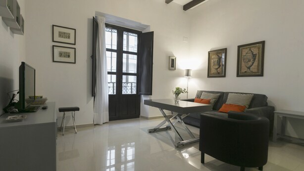 A modern living room with a black sofa, a dining table, and a black bench. There are framed pictures on the walls and a large window with curtains. - Apartamento Estándar (Sevilla)