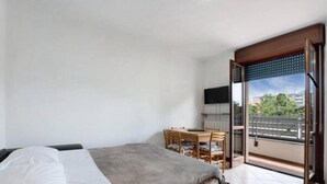 Camera da letto con un letto, una scrivania e una sedia. Sul balcone si può ammirare la vista di edifici e alberi.