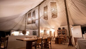 Un espace de restauration aménagé sous une tente, avec des tables et des chaises en bois, des luminaires suspendus et un bar doté d’un buffet en bois.
