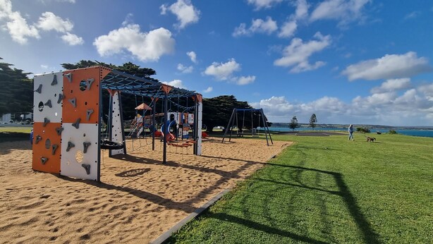 A playground with climbing structures and a swing set, adjacent to a grassy area and a body of water.