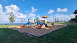 A playground with slides and swings, a sandy play area, and a grassy field with a view of the sea.
