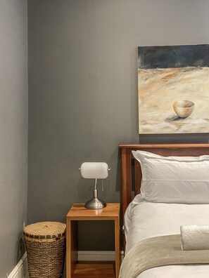 Een slaapkamer met een houten hoofdeinde, een nachtkastje met een lamp, een rieten mand en een schilderij aan de muur.