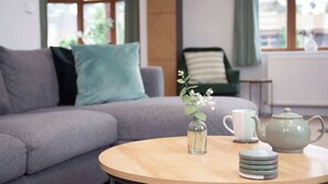Ein modernes Wohnzimmer mit einem grauen Sofa, einem runden Holztisch, einer klaren Glasvase mit Blumen, einer weißen Tasse und einem grünen Teekessel.