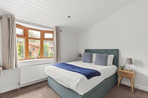 Une chambre avec une grande fenêtre, un lit recouvert d’une couverture bleue et une table de chevet avec une lampe.