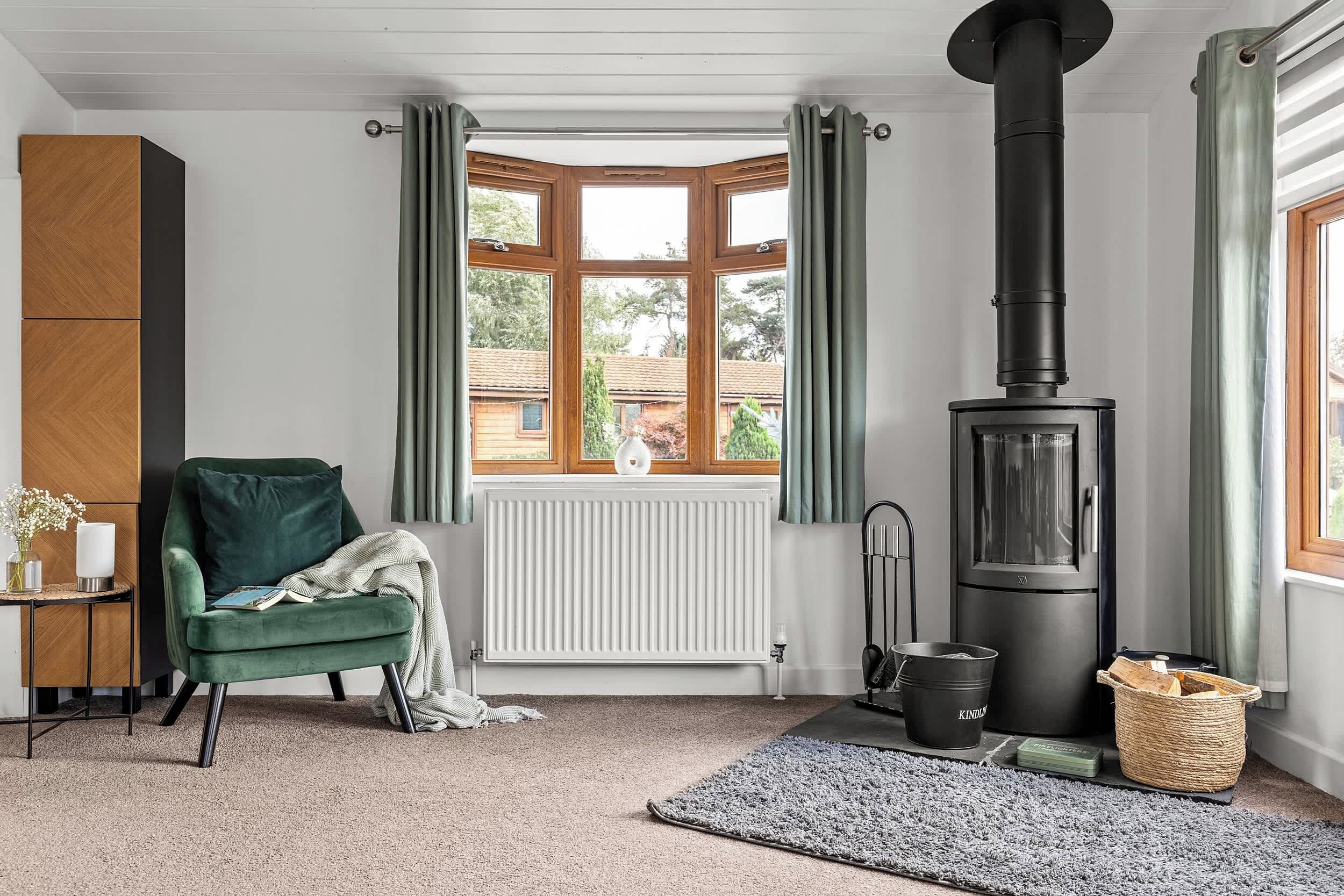 Een gezellige kamer met een groene fauteuil, een witte radiator, een zwarte kachel en uitzicht naar buiten door een raam met groene gordijnen.