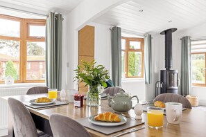 Une table de petit-déjeuner dressée avec des croissants, du jus de fruits, de la confiture et une théière, dans une pièce lumineuse dotée de grandes fenêtres et d’un poêle à bois.