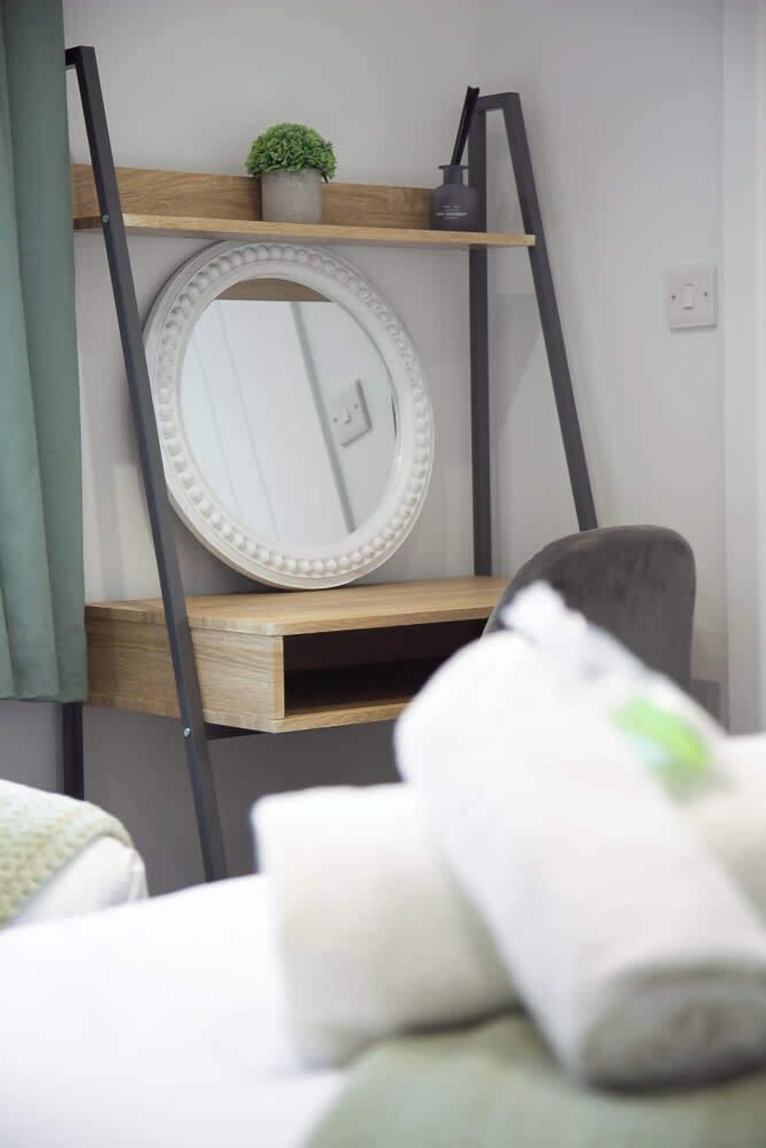 A room with a round mirror on a wooden shelf, a small potted plant, and a rolled-up towel on a table.