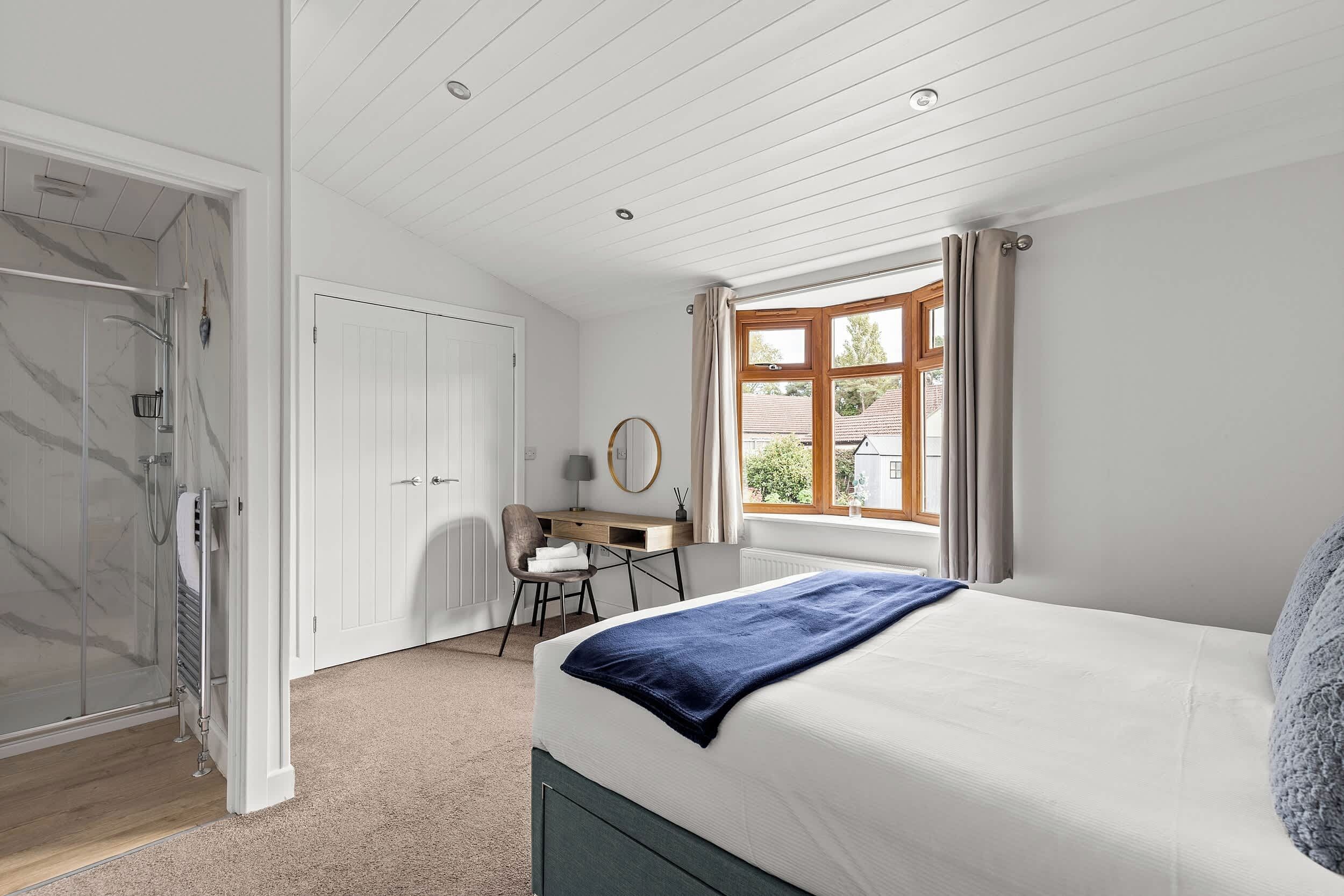 A bedroom with a bed, a desk, a chair, a window, and a door leading to a bathroom.