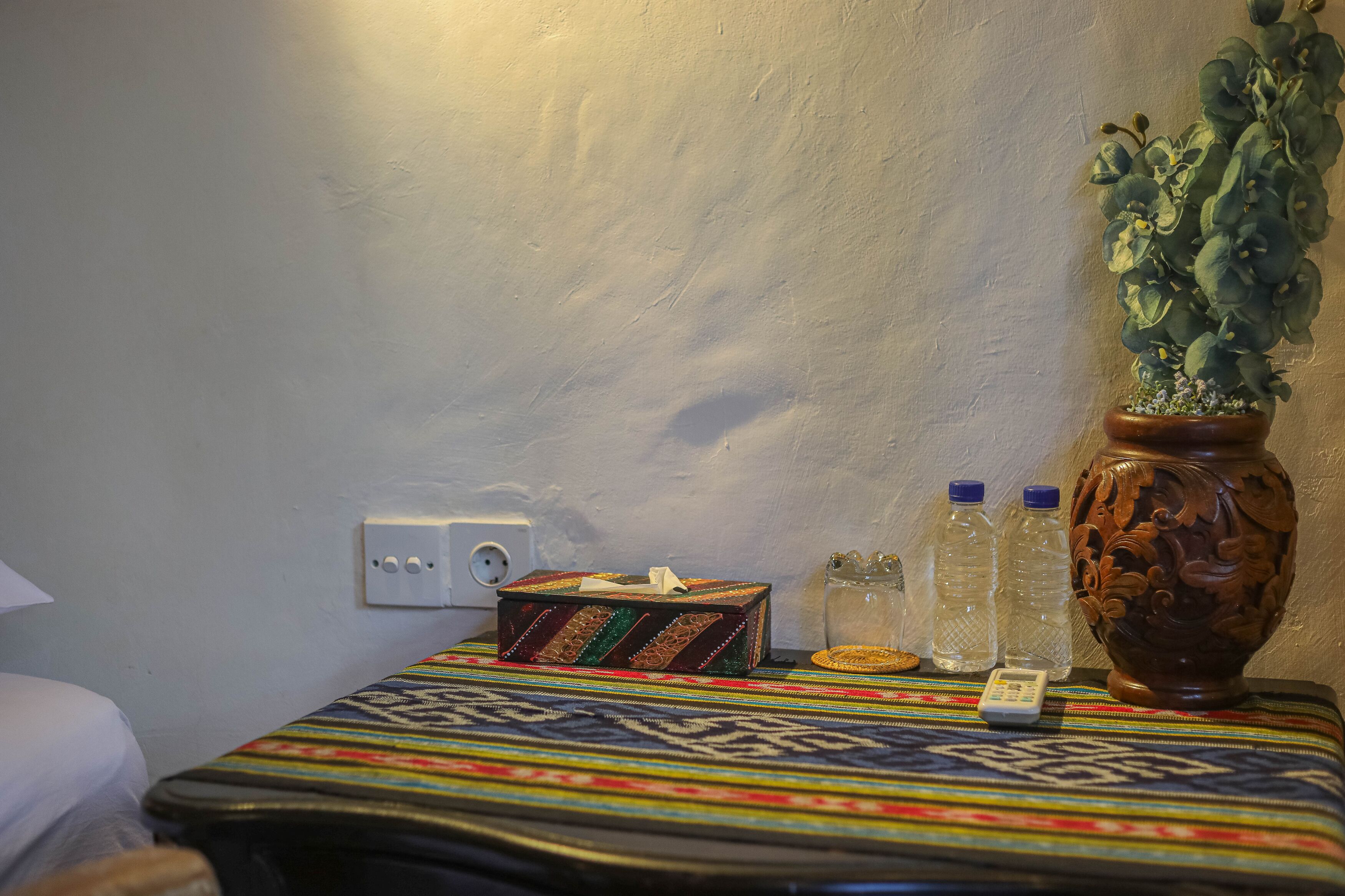 Ein Bett mit farbenfroher Decke, eine kleine dekorative Schachtel, zwei Wasserflaschen, eine Fernbedienung und ein Blumentopf auf einem Nachttisch.