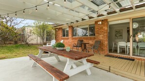 A covered patio with a wooden table and benches, brick walls, and a glass door leading to an indoor dining area. - Berry Haven by Experience Jervis Bay (Vincentia)