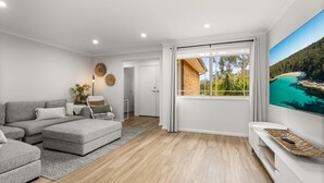 A modern living room with a grey sectional sofa, a white TV stand, and a large window offering a view of a beach and forest. - Berry Haven by Experience Jervis Bay (Vincentia)