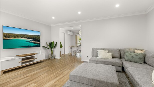 A modern living room with a flat-screen TV, a grey sofa set, a dining area with a white table and chairs, and a kitchen in the background. - Berry Haven by Experience Jervis Bay (Vincentia)