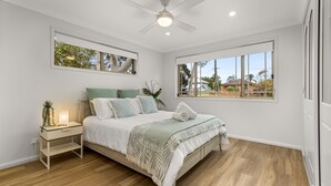 A bedroom with a bed, bedside table, nightstand, and a window with a view of houses and trees. - Berry Haven by Experience Jervis Bay (Vincentia)