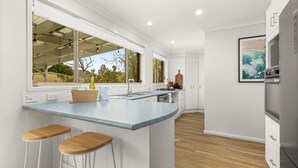 A modern kitchen with a large island, bar stools, a built-in oven, and a view of the outdoors. - Berry Haven by Experience Jervis Bay (Vincentia)