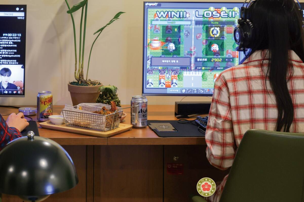Une personne est assise à un bureau, devant un ordinateur dont l’écran affiche un jeu vidéo. On peut également voir une plante, une canette de soda et un téléphone intelligent.