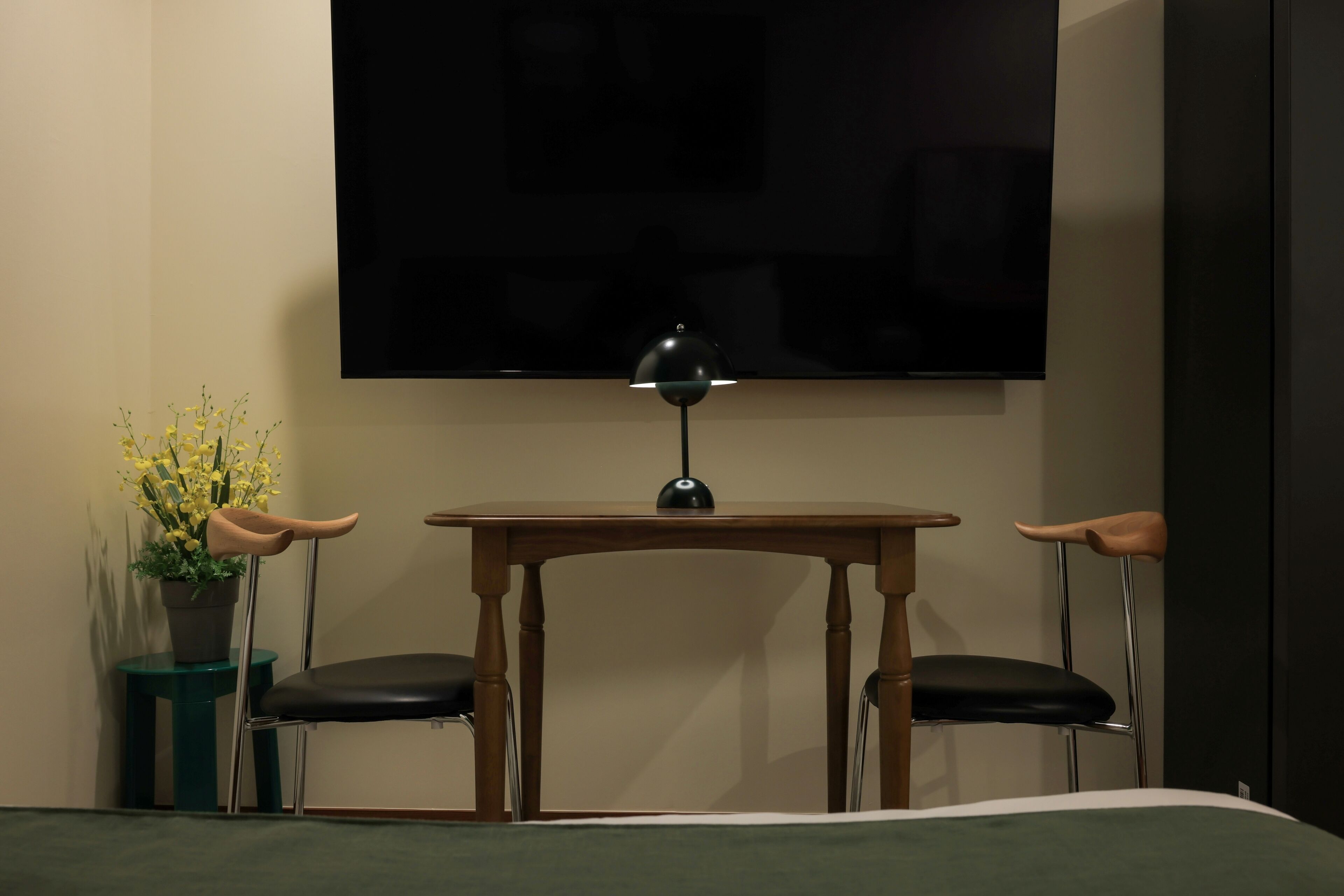 A hotel room with a bed, a TV mounted on the wall, a small wooden table with two chairs, and a potted plant.