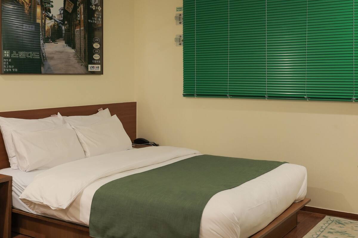 Un lit bien fait, avec une literie blanche et un plaid vert, une table de chevet avec une horloge et des mouchoirs, une petite poubelle et un store vert à la fenêtre.