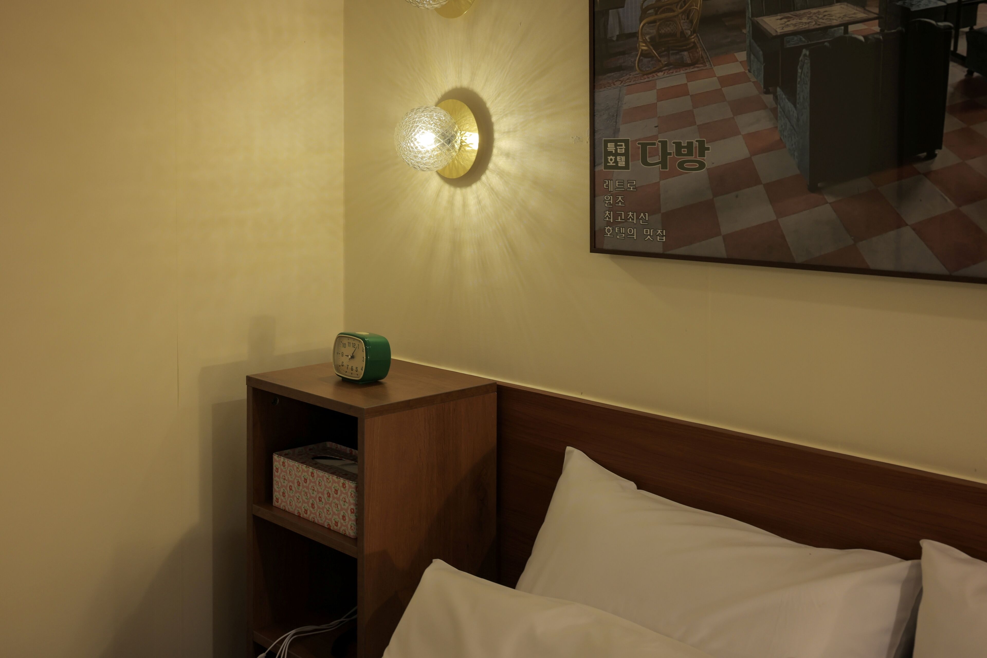 Een hotelkamer met een houten hoofdeinde, een bed met witte lakens, een nachtkastje met een groene wekker en een wandlamp.