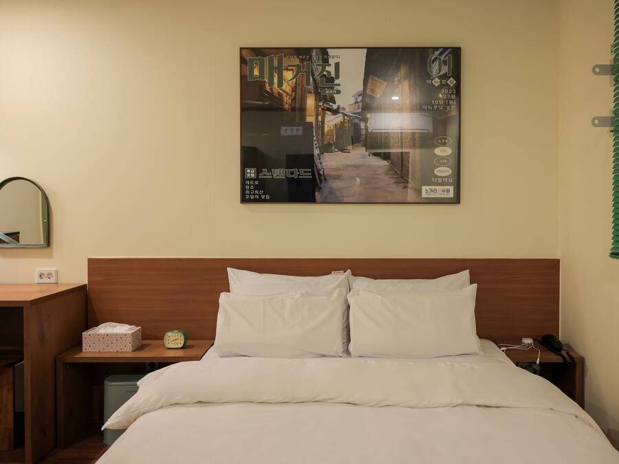 A hotel room with a neatly made bed, a bedside table with a lamp, a framed picture on the wall, and a window with blinds.