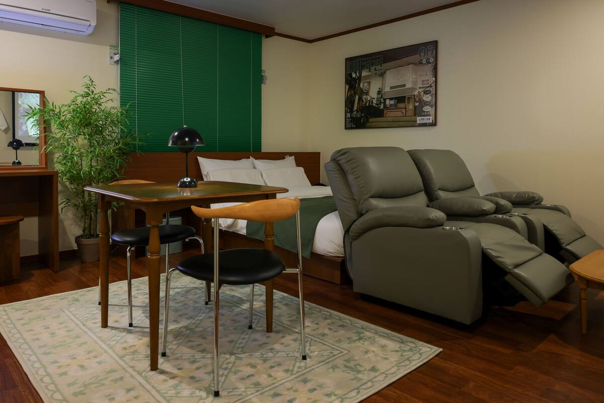 Une pièce de taille modeste comprenant un lit, un fauteuil massant, une petite table avec des chaises, une plante en pot et un climatiseur fixé au mur.