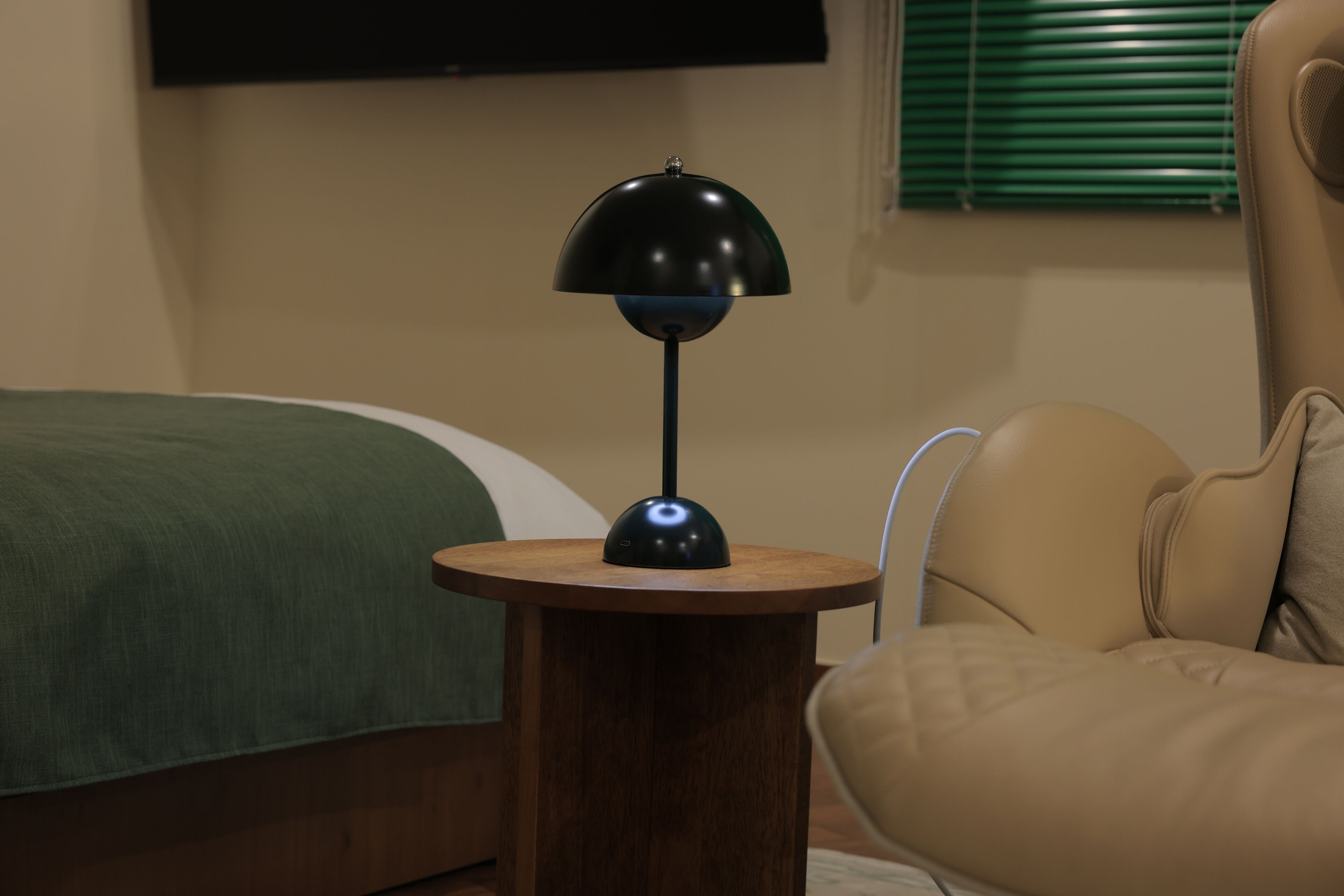 Una mesa redonda de madera con una lámpara negra, una cama con una manta verde y un sillón color beige.