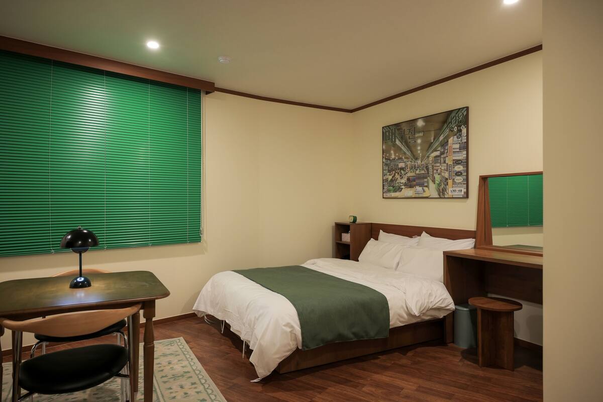 Une chambre à coucher comprenant un lit, un bureau, une chaise, un petit tabouret et une fenêtre avec des stores.