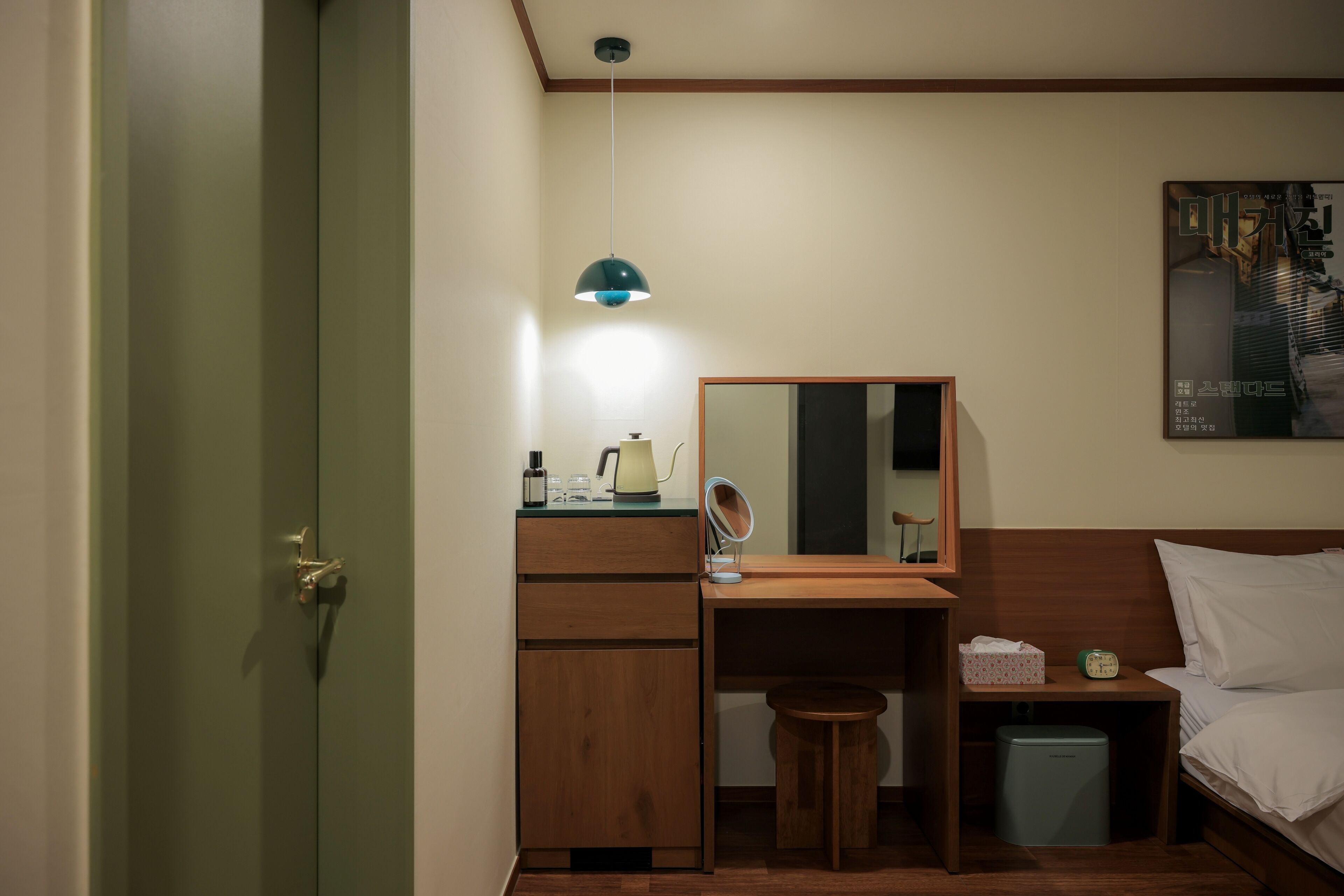 Habitación de hotel compacta con escritorio de madera, taburete pequeño, mesita de noche con lámpara, espejo, cama y cubo de basura.