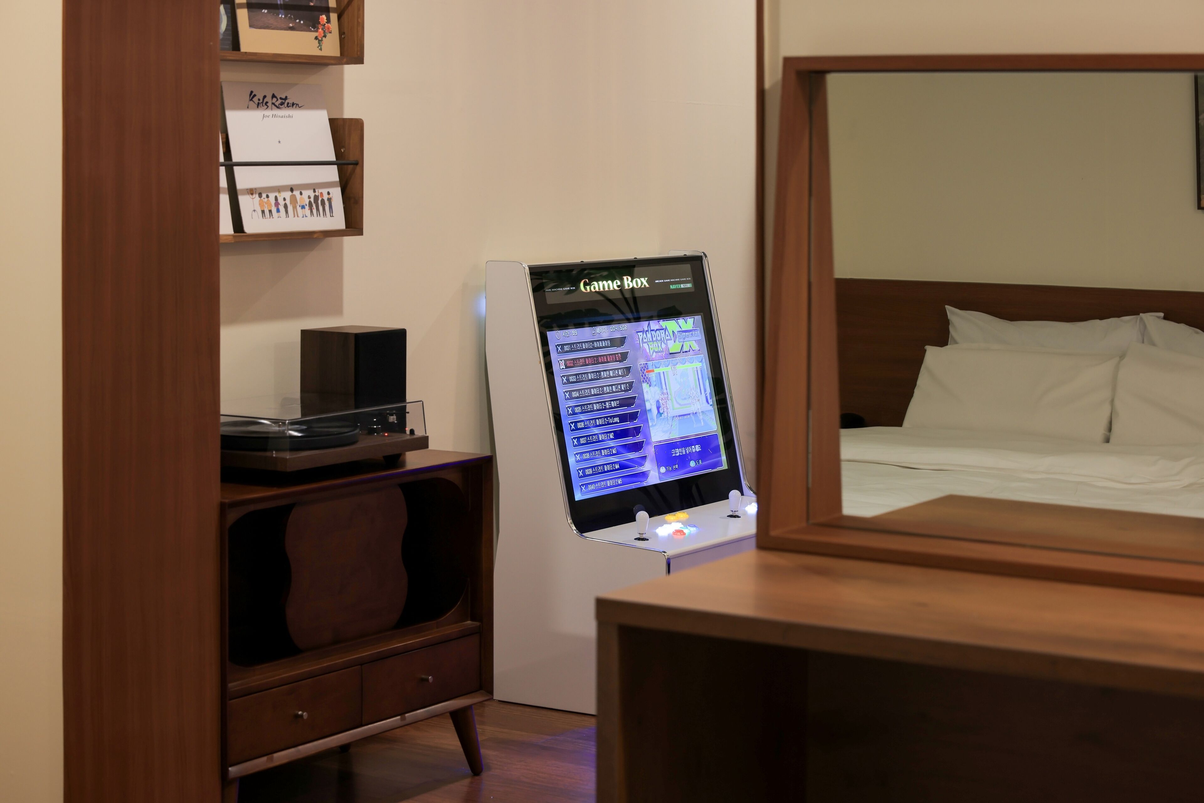 A hotel room with a bed, a wooden nightstand, a GameBox, and a shelf with items.