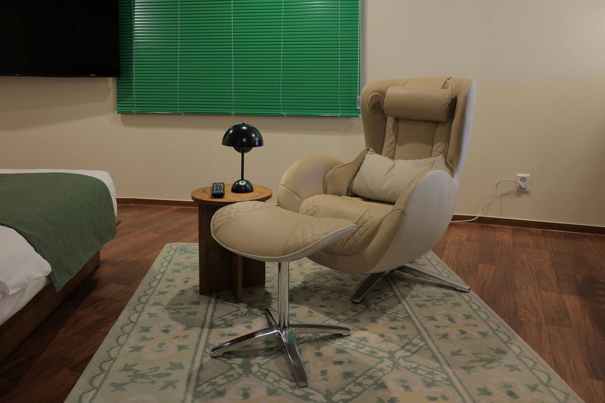 Une chambre moderne avec un fauteuil beige, une petite table d’appoint en bois sur laquelle se trouve une lampe, un tapis à motifs verts et un lit recouvert d’une couverture verte.