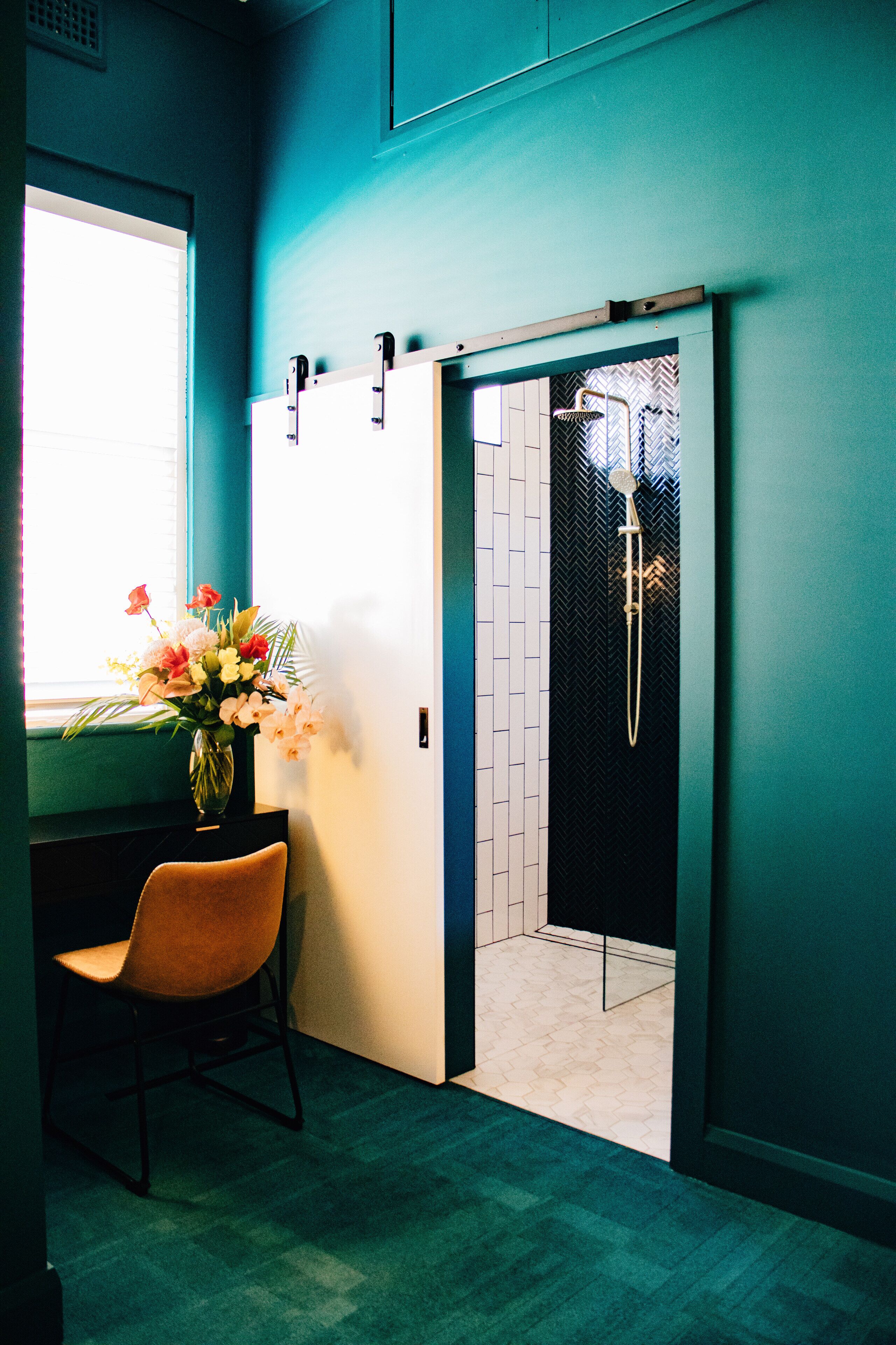 Een badkamer met een douche, een raam en een vaas met bloemen op een bureau.