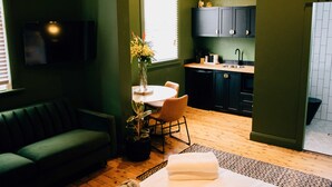 A compact living space with a green wall, a sofa, a dining table with chairs, a small kitchen area, and a TV mounted on the wall.