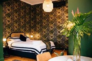 A bedroom with a patterned wallpaper, a bed with white bedding, a bedside table with a lamp, and a potted plant. - Rooms of Renown (Leeton)
