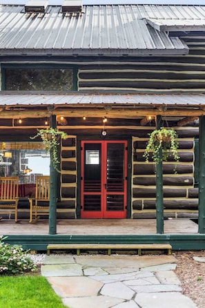 A log cabin with a red door, a porch with hanging plants, and a stone pathway leading to the entrance.