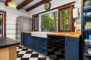 A kitchen with a black and white checkered floor, wooden beams on the ceiling, a stainless steel refrigerator, and a window with a view of trees.