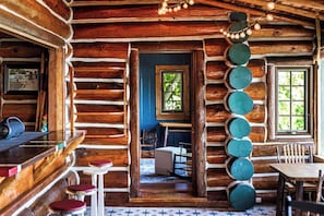 A rustic log cabin interior with a dining area, a view through a doorway, and a decorative light fixture.
