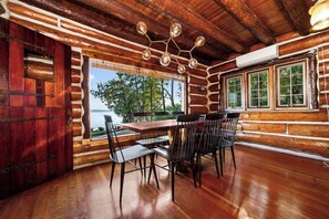A log cabin interior with a wooden dining table and chairs, a large window with a view of trees and water, and a rustic ceiling with exposed beams. - Paradesia Cove (Northport)