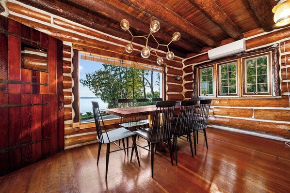 A log cabin interior with a wooden dining table and chairs, a large window with a view of trees and water, and a rustic ceiling with exposed beams. - Paradesia Cove (Northport)