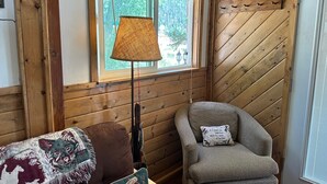 Ein gemütliches Zimmer mit Holzwänden, einem Fenster mit Jalousien, einer Lampe, einem Sessel mit Kissen und einer gemusterten Decke.
