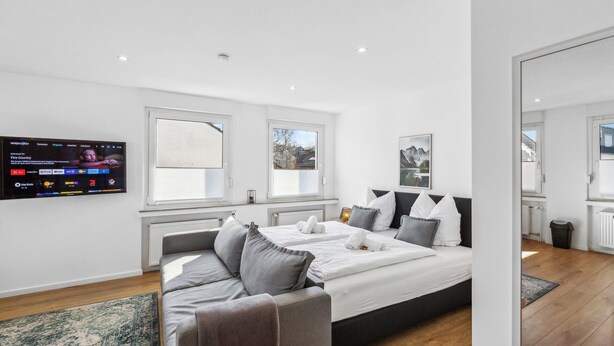 A modern bedroom with a large bed, a grey sofa, and a TV mounted on the wall displaying various channels.