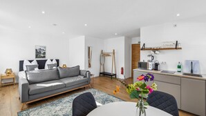 A modern living room with a grey sofa, a dining table with a vase of flowers, and a kitchen area with a microwave, kettle, and shelves.