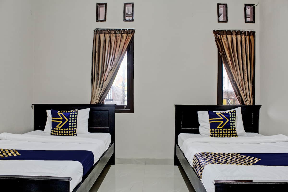Une chambre d’hôtel avec deux lits simples, chacun doté d’une tête de lit, de draps blancs et d’un oreiller bleu et jaune. Il y a deux fenêtres avec des rideaux, et les murs sont de couleur unie, ornés de quelques tableaux encadrés.
