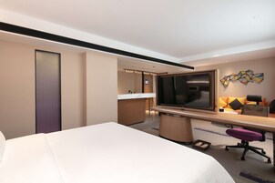 Habitación de hotel con una cama grande, un escritorio con televisor, una kitchenette pequeña y un baño con ducha e inodoro.
