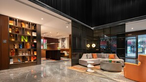 Een moderne lobby met een marmeren vloer, een zithoek met een bank en fauteuils, een boekenkast met verlichte letters, en een groot raam met uitzicht op de stad.