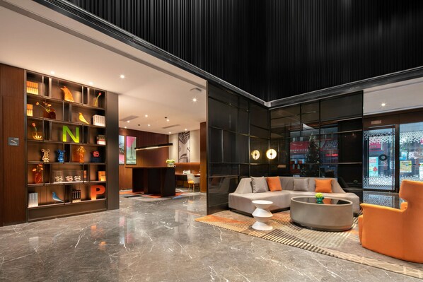 A modern lobby with a marble floor, a seating area with a sofa and armchairs, a bookshelf with illuminated letters, and a large window with a view of the city.