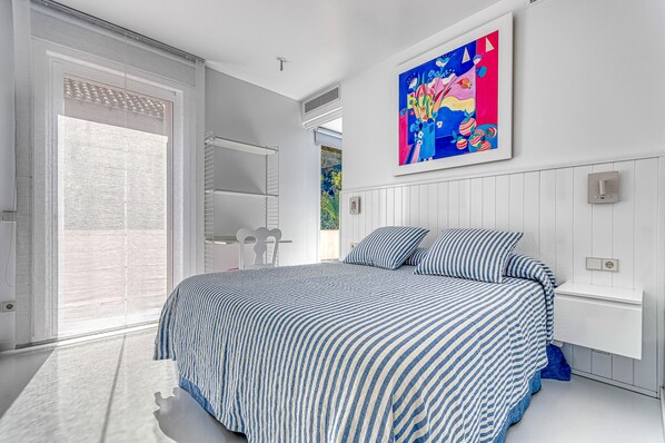 A bedroom with a bed, a large window, a painting on the wall, and a white bedside table. - The Pink Elephant Hotel (Sant Feliu de Guixols)