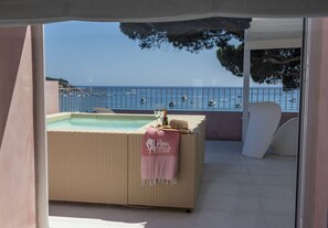 A balcony with a wicker sofa, a small pool, and a view of boats on the water. - The Pink Elephant Hotel (Sant Feliu de Guixols)