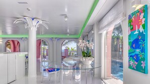 Un interior moderno con una mesa de comedor de vidrio, sillas transparentes, columnas blancas y un cuadro de colores en la pared.