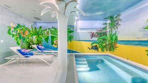 Una piscina interior con un mural tropical, sillas de descanso y un techo con detalles decorativos.